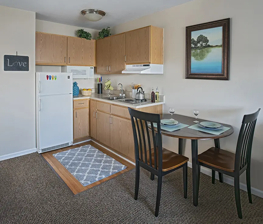 Kitchenette with small dining table and a landscape painting of a pond with trees at West Bloomfield senior housing